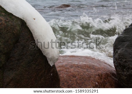 Baltic sea water with waves between two stones covered with snow. The wave is breaking on stones and flying spray