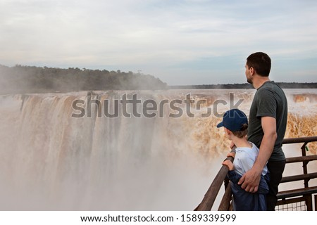 family of two, father and son, enjoying the gorgeous view of devil's throat waterfall (garganta del diablo) at iguazu falls in argentina and brazil, adventure vacation concept Royalty-Free Stock Photo #1589339599
