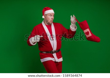 Emotional male actor in a costume of Santa Claus holds a Christmas sock for gifts in his hands and poses on a green chrome background