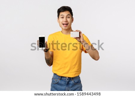 Happy smiling, cheerful asian guy in yellow t-shirt with tattoos, holding smartphone and credit card, recommend using online cashback, finance system, order product internet, white background