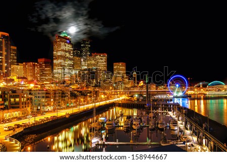 Seattle night view of the illuminated waterfront and skyline under a full moon breaking through clouds