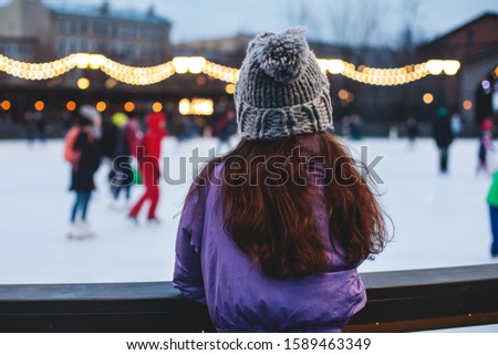 Girl watching on the ice rink with happy people ice skating in the background, concept of ice skating in winter, holiday christmas time, with new year decoration and illumination, Helsinki, Finland