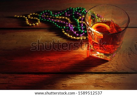 Close-up view of sazerac cocktail with a lemon peel in glass on wooden table with side lighting and warm tones. Mardi gras beads in green, gold and purple nearby.