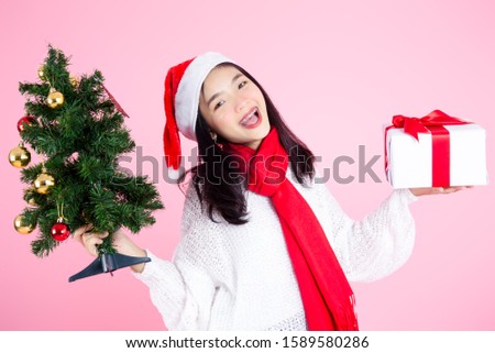 Asian teenager girl wearing Santa hat holding Christmas tree and gift box on pink.