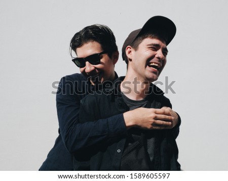 Two funny guys hugs and laugh in dark clothes at white wall background sunny day 