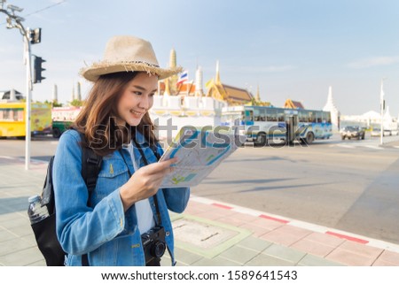 Beautiful smile Asian woman tourist in a jean jacket and straw hat, she watching a map on her hand searching for attraction place in Bangkok for travel and rest with Wat Phra Kaew background.