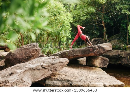 Pretty young slim woman doing one-legged upward facing dog pose when working out in forest