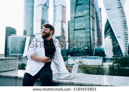 Happy content  bearded man in glasses dressed in white shirt texting on mobile phone standing near embankment in city with skyscrapers 