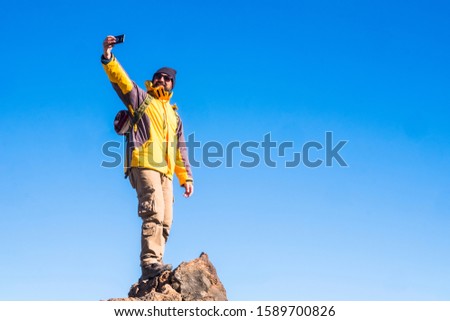 Adventure man standing outdoor on the top of a mountain with blue sky in background - concept of travel and trekking or hiking sport activity - people and technology outdoor