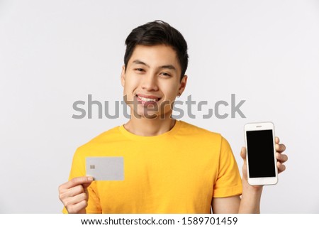 Handsome and cute smiling asian man in yellow t-shirt, showing smartphone display and credit card, grinning delighted, recommend bank service, online paying system, standing white background