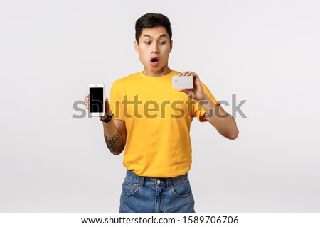 Impressed and surprised good-looking asian, chinese hipster guy in yellow t-shirt, holding smartphone and credit card, stare at banking card with wow face, gasping astonished, white background