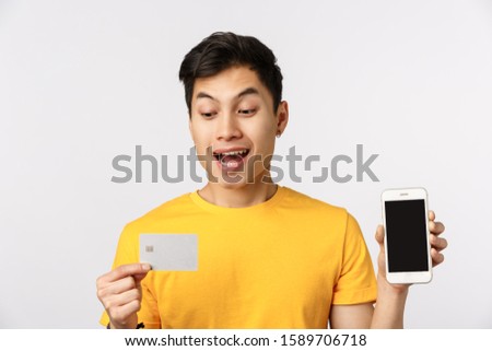 Close-up shot enthusiastic guy decided put money deposit, get credit to pay for trip, holding smartphone showing display and banking card, smiling amused, standing white background