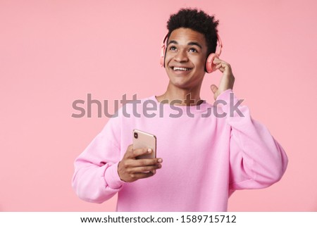 Portrait of a smiling african teenager boy wearing pullower standing isolated over pink background, listening to music with headphones, holding mobile phone, dancing