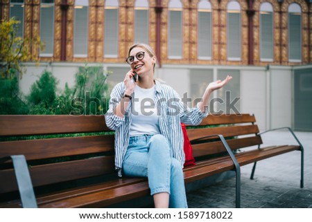 Trendy blonde woman in casual wear with eyeglasses sitting on wooden bench with crossed legs gesticulating and speaking on phone