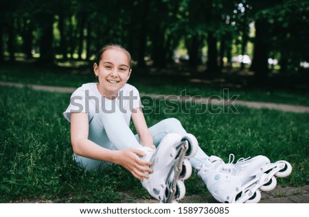 Joyful and delighted child sitting on lawn in park and putting on roller skates while smiling and looking at camera