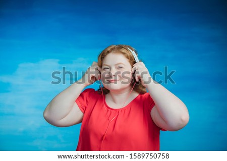Young attractive plump woman with red hair listening to music with pleasure headphones on a blue background.