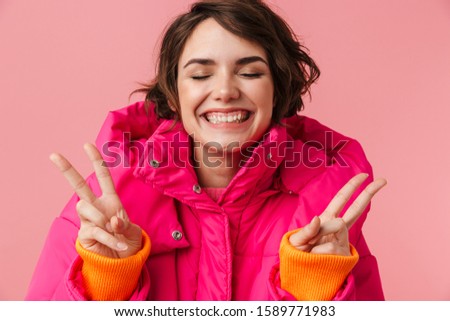 Portrait of young happy woman in warm coat with gesturing peace sign and smiling isolated over pink background