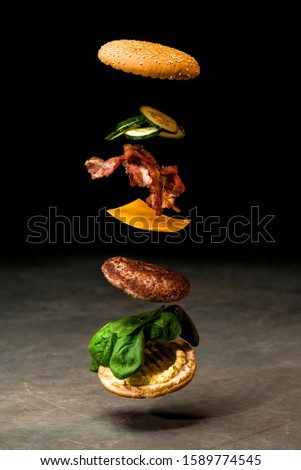 Flying cheeseburger ingredients on dark background of concrete. Fastfood concept. Ingredients of burger - bun, grilled cutlet, cheese, bacon, cucumber and spinach Royalty-Free Stock Photo #1589774545