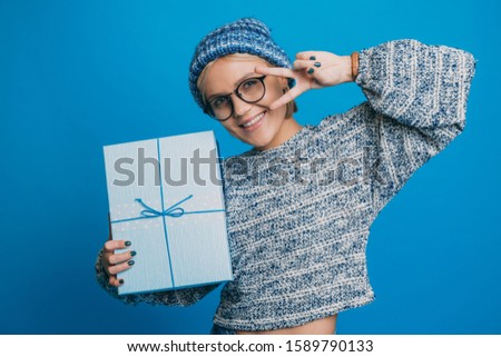 Portrait of a charming young woman holding a blue gift box present while looking at camera smiling against blue background.