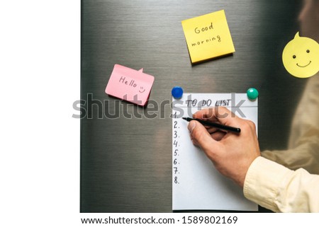 cropped view of man filling in to do list hanging on fridge near wishes on sticky notes isolated on white