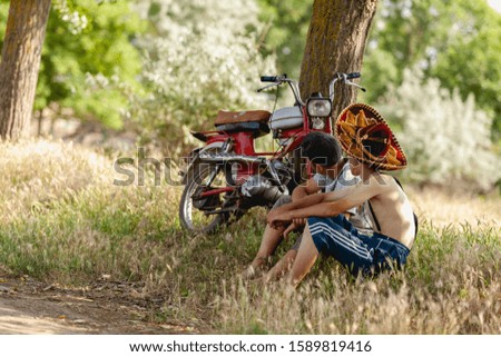 Two boys communicate sitting on the grass near the retro motorcycle. Boy in a sombrero resting after cycling. Summer activity concept.