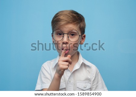 Funny little caucasian boy in glasses shows a sign of silence bowing his finger over a card on a blue background. Concept of personal offers and promotions in children's stores. Advertising space