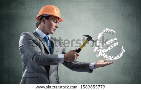 Businessman going to crash with hammer euro currency sign. Young handsome man in business suit and safety helmet standing on grey wall background. Financial crisis, bankruptcy and inflation pressure.