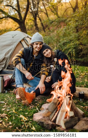 Young loving couple of tourists relaxing near the fire in the nature.
Handsome man and beautiful woman are roasting marshmallows over the fire near tent in camping. Instagram stories picture.