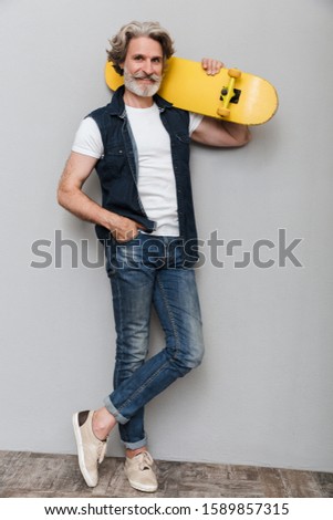Full length portrait of a handsome stylish mature man wearing a vest standing over gray background, posing with a skateboard on his shoulder