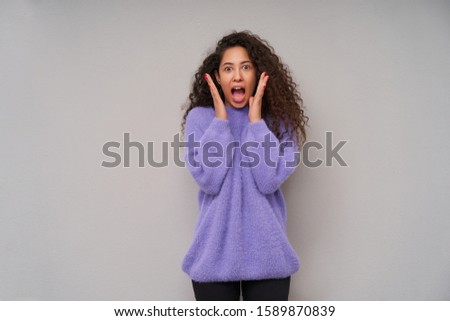 Portrait of excited young pretty curly brunette lady raising amazedly palms to her head and rounding eyes while looking at camera, posing over grey background in casual clothes