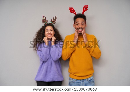 Indoor photo of happy young dark haired curly pair keeping hands on their faces and smiling excitedly at camera, showing their pleasant emotions while standing over grey background