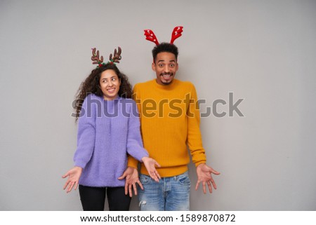 Cheerful young lovely dark skinned curly pair wearing holiday hoops while preparing for christmas party, smiling positively and shrugging with opened arms over grey background
