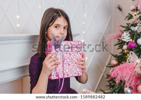 a seven-year-old girl with black hair in a purple dress holds a gift box in her hands near a Christmas tree indoors. pink feathers on the Christmas tree.Christmas gift