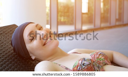 Portrait of young woman in cap is resting laying on deckchair after swimming in pool. She looking at camera and smiling.