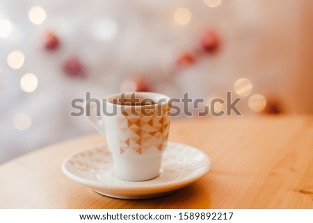 Cup of espresso or americano coffee in white cup in cosy Christmas arrangement with festive lights decoration with golden bokeh background, copy space