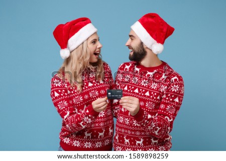 Cheerful young couple guy girl in Christmas sweater Santa hat posing isolated on blue wall background. Happy New Year 2020 celebration holiday party concept. Mock up copy space. Hold credit bank card