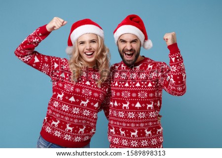 Happy joyful young couple guy girl in Christmas sweaters, Santa hat posing isolated on blue wall background. New Year 2020 celebration holiday party concept. Mock up copy space. Doing winner gesture