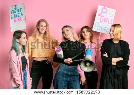 independence, feminism, freedom concept. five young girls feminists advocating feminism isolated over pink background, using megaphone or loud speaker and posters, propaganda for society