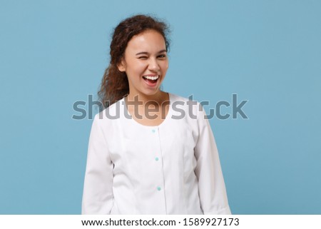 Cheerful african american doctor woman isolated on blue wall background studio portrait. Female doctor in white medical gown blinking. Healthcare personnel health medicine concept. Mock up copy space