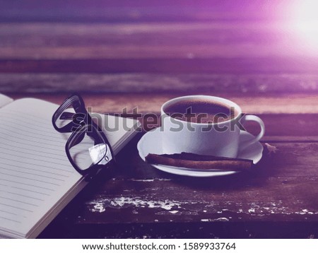 Coffee cup has Americano coffee,blurred book and eyeglasses on old wood table top,with lens flare,free space for tour text design,top view.Concept of Relaxation in Coffee time.