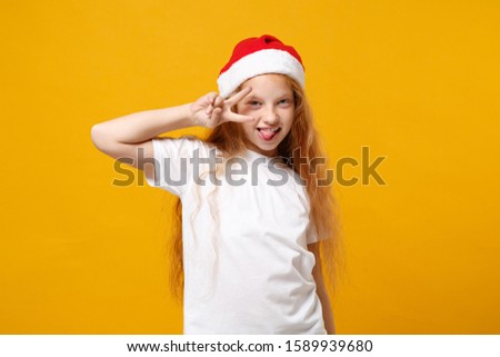 Funny little ginger kid Santa girl 12-13 years old in white t-shirt Christmas hat isolated on yellow background. New Year 2020 celebration holiday concept. Mock up copy space. Showing victory sign