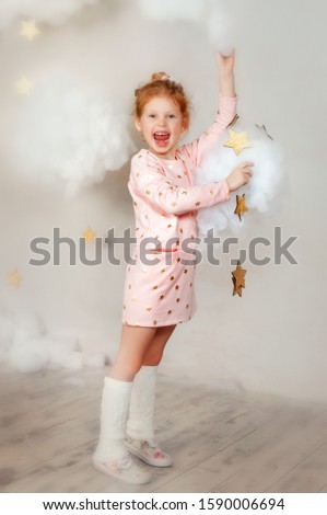5 years old girl in pink dress with star print on it and white socks stay in clouds and stars with wide smile. Beautiful little girl touch stars and sky. Smiling dreamy child dream about magic.
