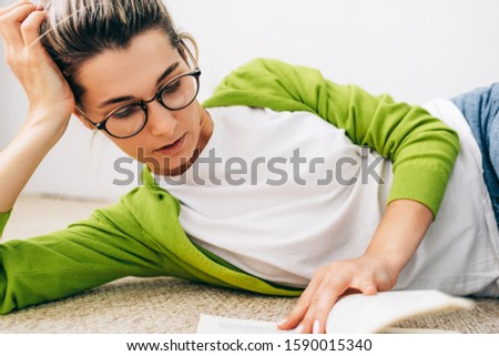 Close-up image of smart young woman reading book, wearing green cardigan and white t-shirt, eyeglasses, lying on the carpet. Female relaxing during reading a book. Student girl studying at home.