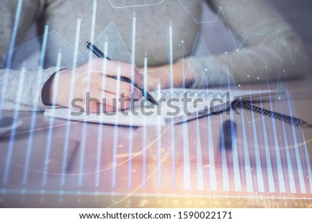 Hands writing information about stock market in notepad. Forex chart holograms in front. Concept of research. Multi exposure