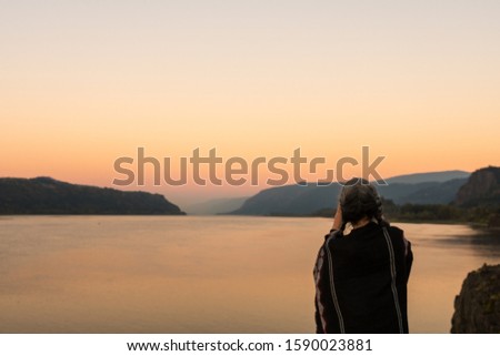 A young woman with gray hair and a poncho takes photographs of the sunset at the Corbett lookout on the Columbia River, Oregon, USA