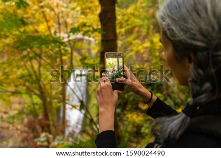A young woman with gray hair takes photographs of the Multnomah waterfall at Multnomah Creek in the Columbia River Gorge, Oregon, USA