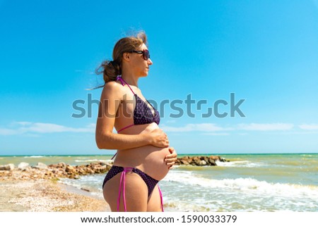 A pregnant girl holds a big belly with her hands on the beach by the sea.