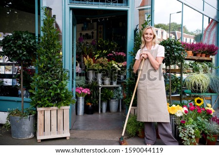 Woman florist standing at the entrance to her shop, leaning on a broom Royalty-Free Stock Photo #1590044119
