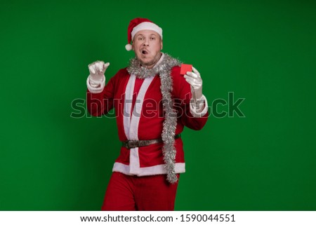 Emotional male actor in a costume of Santa Claus holds a small red gift box in his hands and poses on a green chrome background