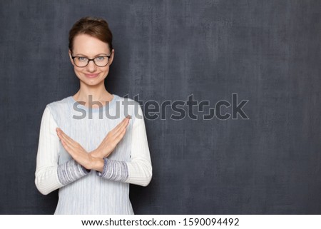 Studio half-length portrait of happy girl with glasses, wearing jumper, making cross with hands, showing stop or prohibition gesture, rejecting something, over gray background, copy space on right